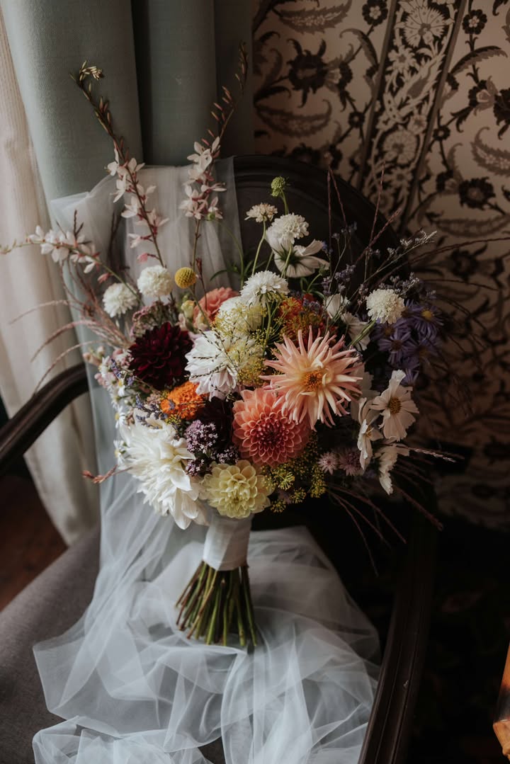 A wedding bouquet arranged by WilderBlooms sustainable floristry sits on top of a bridal veil on a chair. The bouquet is made of british grown sustainable flowers