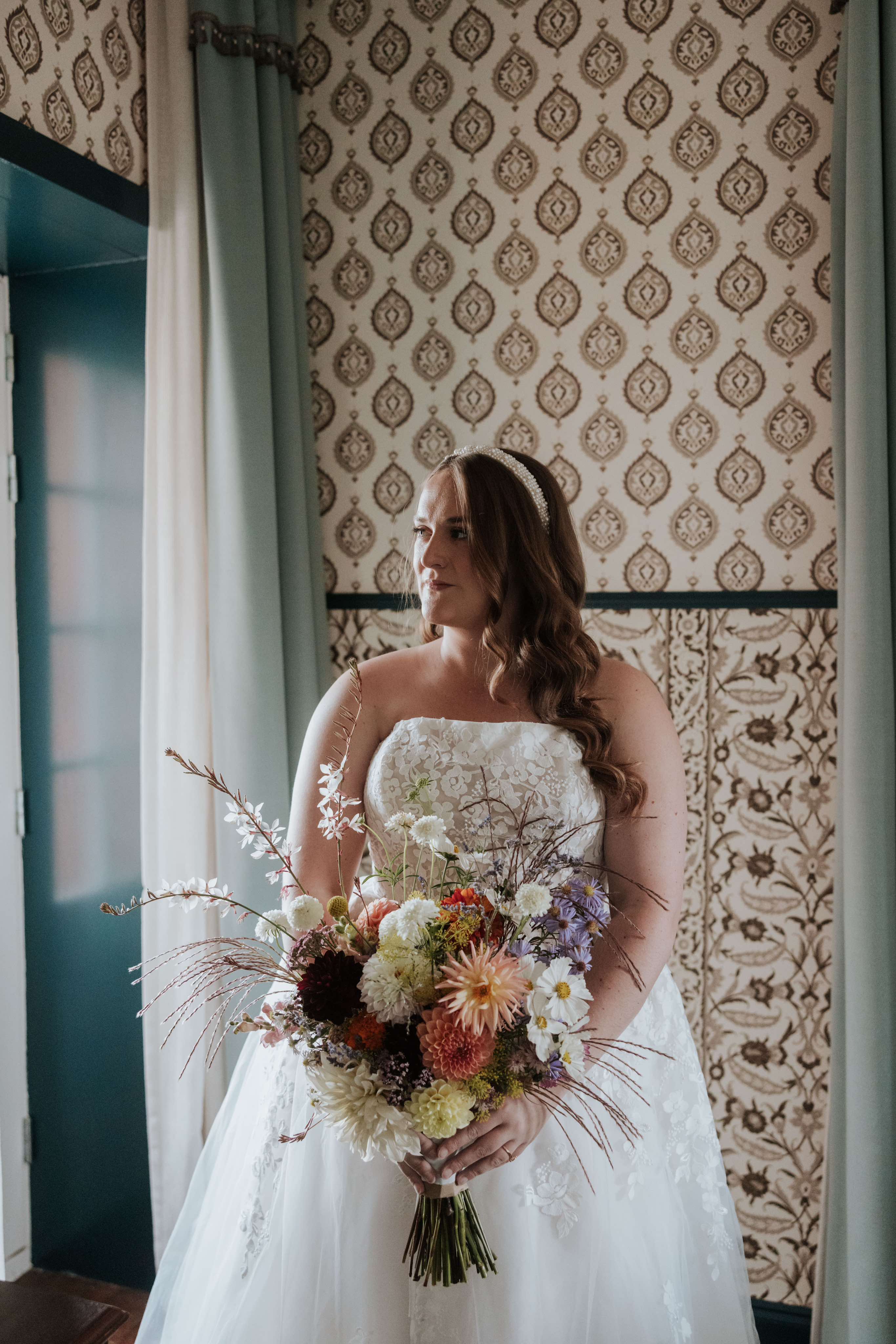 bride stands in a hotel room holding a large wild bouquet of sustainable, eco friendly, locally grown flowers. these british grown  bridal flowers are pastel colours and include dahlias and cosmos