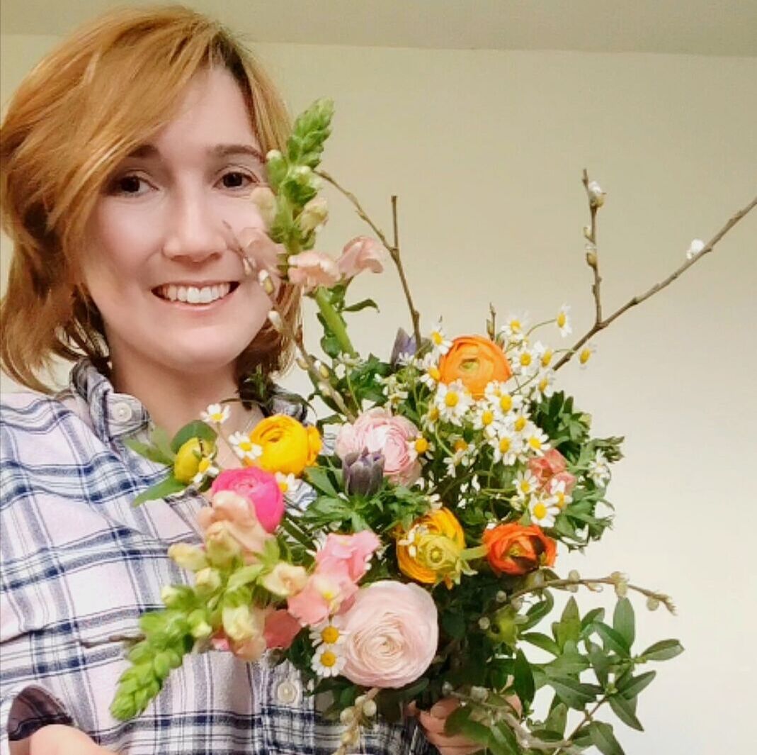 The florist, Emma, smiles and poses with an arrangement of locally grown, sustainable flowers. They are pink, orange, purple and white, with branches and lots of green leaves.